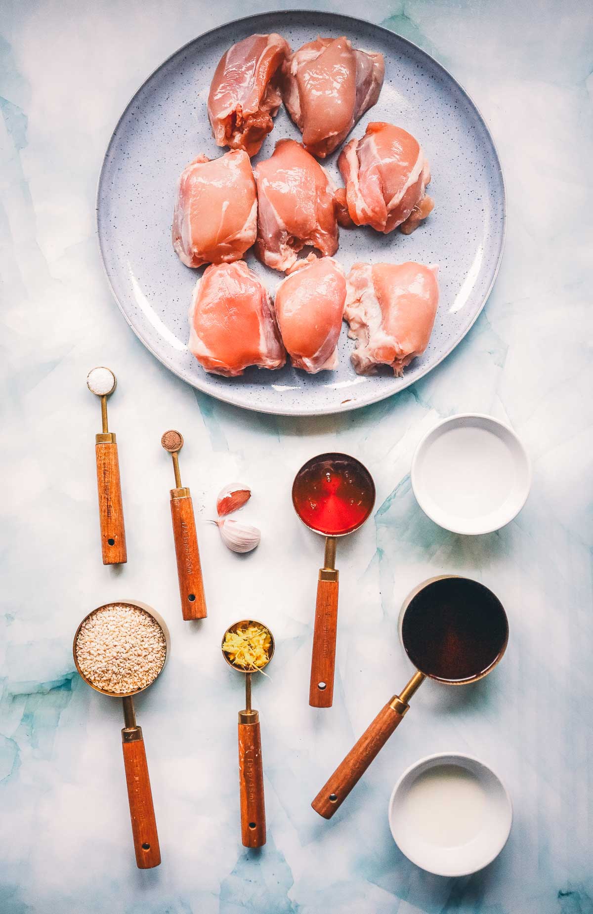 Ingredients for baked teriyaki chicken thighs: raw boneless skinless thighs, soy sauce, sesame seeds, grated ginger, garlic cloves and measured liquids.