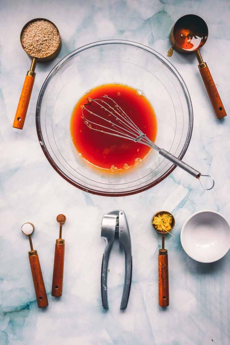 Whisking the soy and honey teriyaki marinade in a glass bowl, with a garlic press, grated ginger and sesame seeds set out beside it.