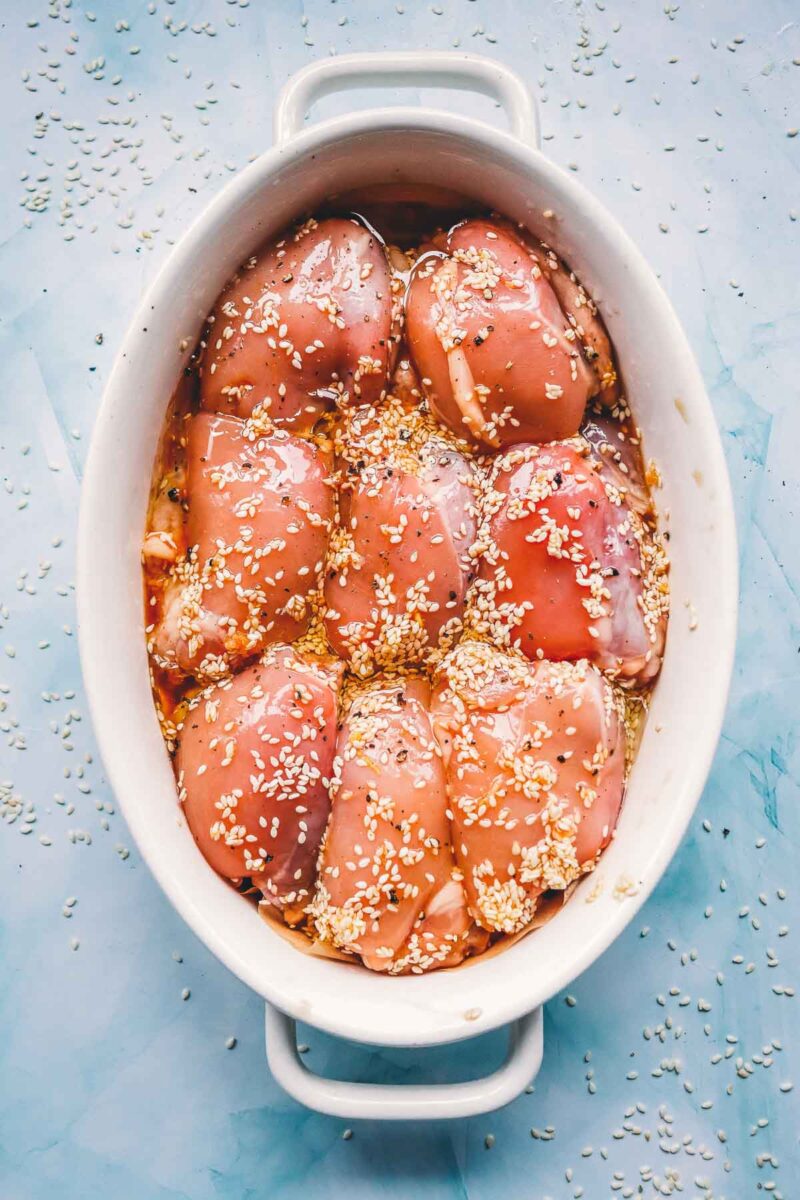 Marinated raw chicken thighs in a white oval baking dish, coated in teriyaki marinade and sprinkled with sesame seeds before baking.
