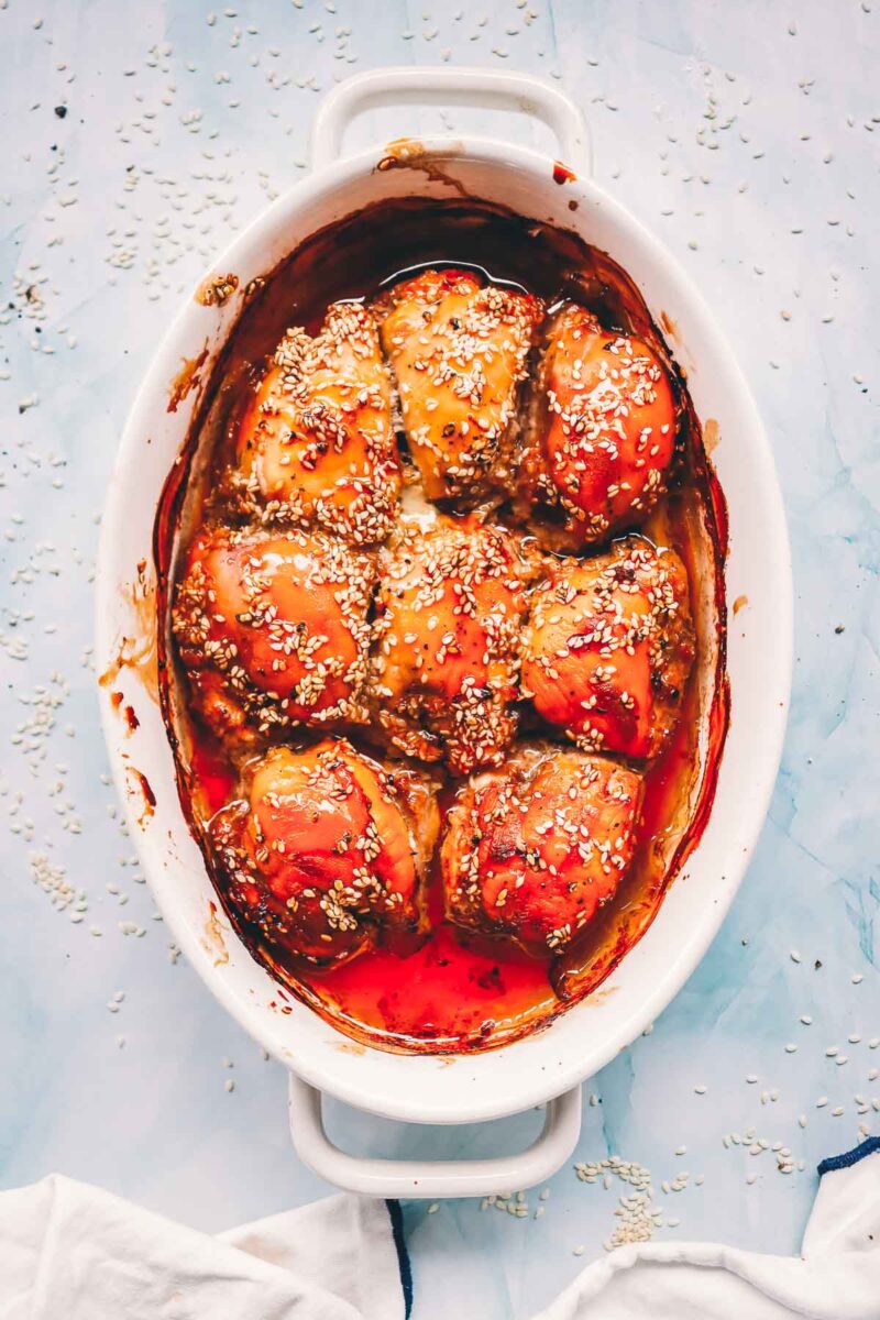 Baked teriyaki chicken thighs straight from the oven in a white oval dish, glazed deep amber with sauce pooled around the edges.