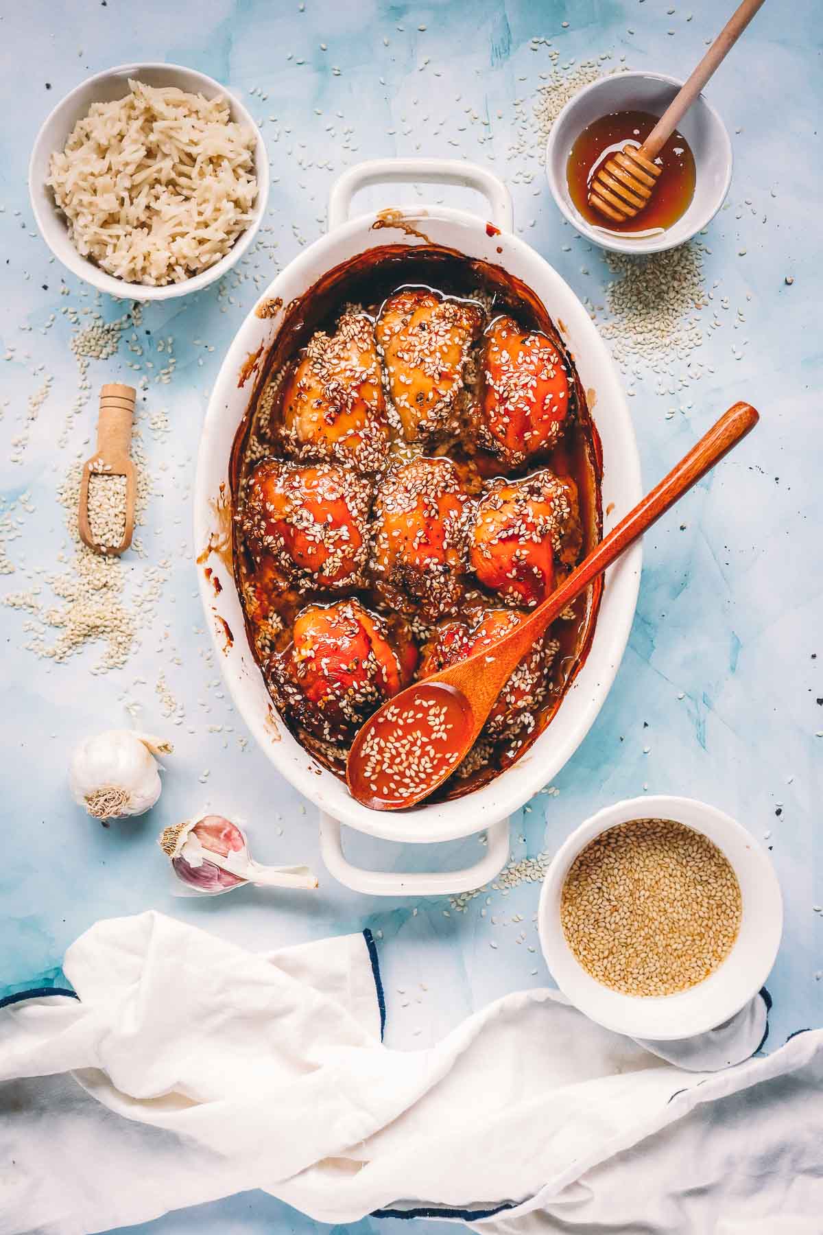 Overhead of baked teriyaki chicken thighs in a white dish with a wooden spoon, set with bowls of white rice, honey, sesame seeds and garlic.