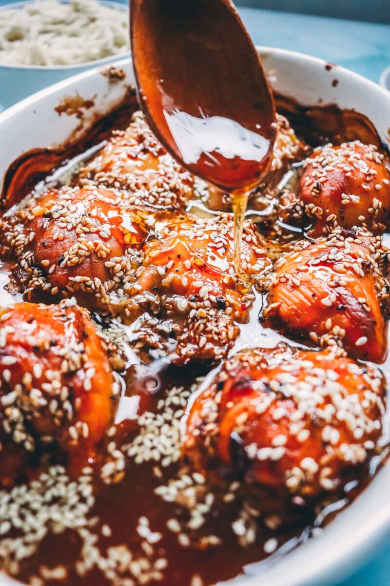 A wooden spoon basting glossy teriyaki pan sauce over sesame-crusted baked chicken thighs in the dish.