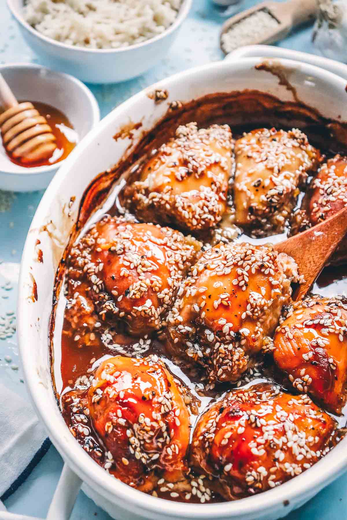 Close-up of baked teriyaki chicken thighs shining with honey soy glaze and sesame seeds, with rice and a honey dipper behind.
