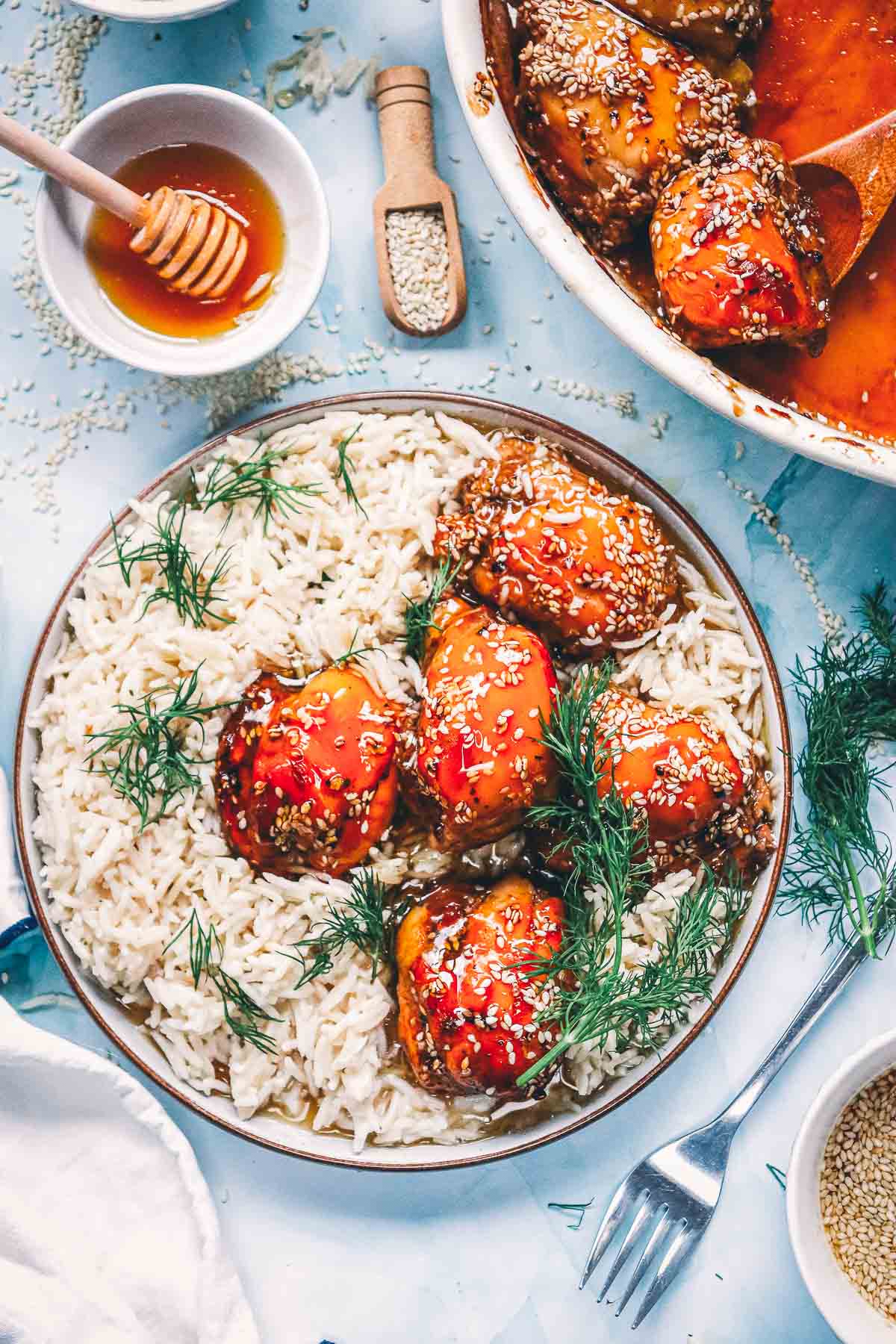 Baked teriyaki chicken thighs served over white rice with fresh dill and sesame seeds, next to the baking dish and a bowl of honey.