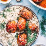 Glazed baked teriyaki chicken thighs over white rice with fresh dill, a fork, a bowl of sesame seeds and honey on the table.