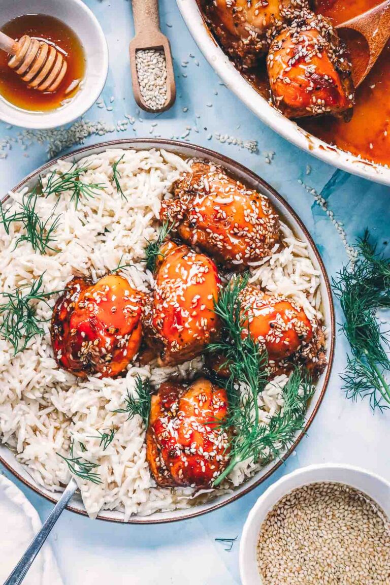 Glazed baked teriyaki chicken thighs over white rice with fresh dill, a fork, a bowl of sesame seeds and honey on the table.