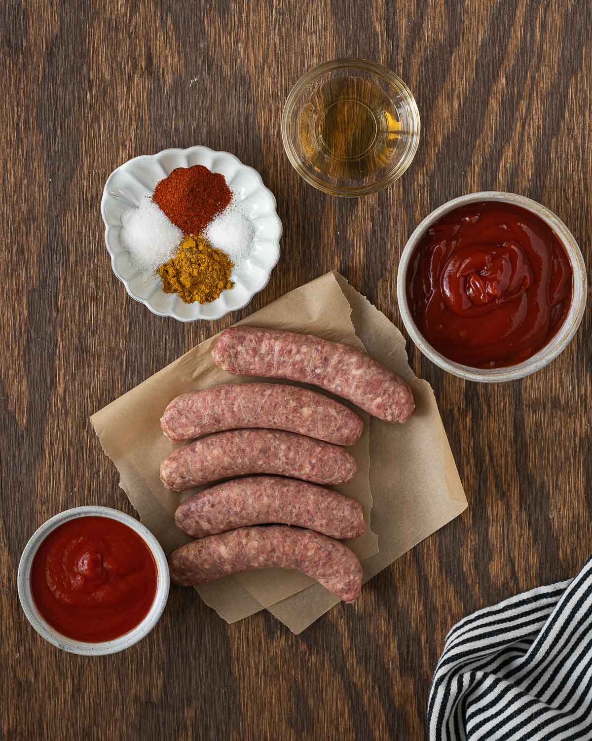 Five raw sausages on parchment paper with bowls of ketchup, spices, and a small glass of liquid, all on a wooden surface next to a striped towel.