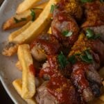 Plate of French fries and sliced sausages topped with curry powder, ketchup, and chopped parsley.
