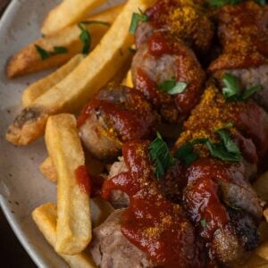 Plate of French fries and sliced sausages topped with curry powder, ketchup, and chopped parsley.