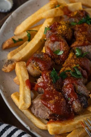 Plate of French fries and sliced sausages topped with curry powder, ketchup, and chopped parsley.