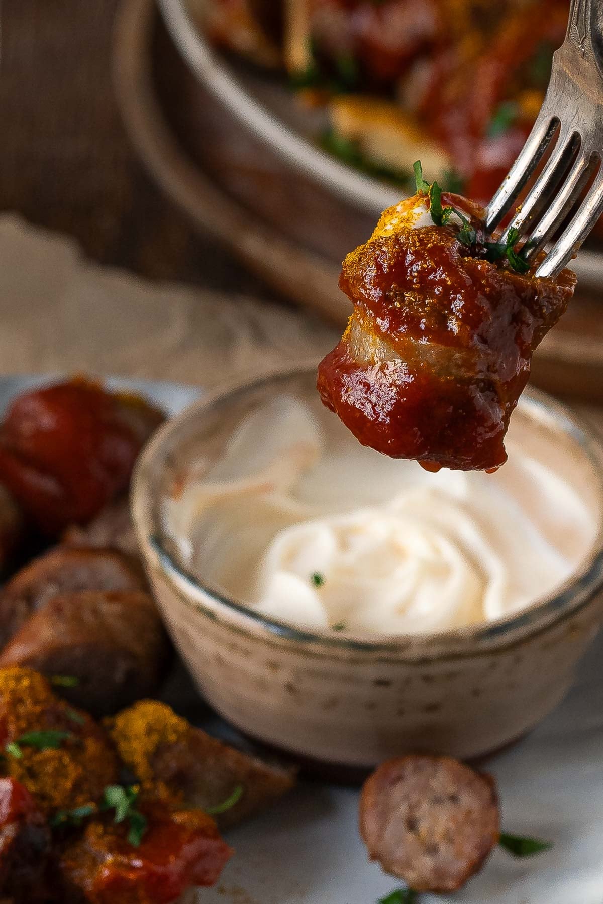 A fork holds a sausage piece with sauce above a bowl of creamy dip, with more sausage pieces and a plate in the background.