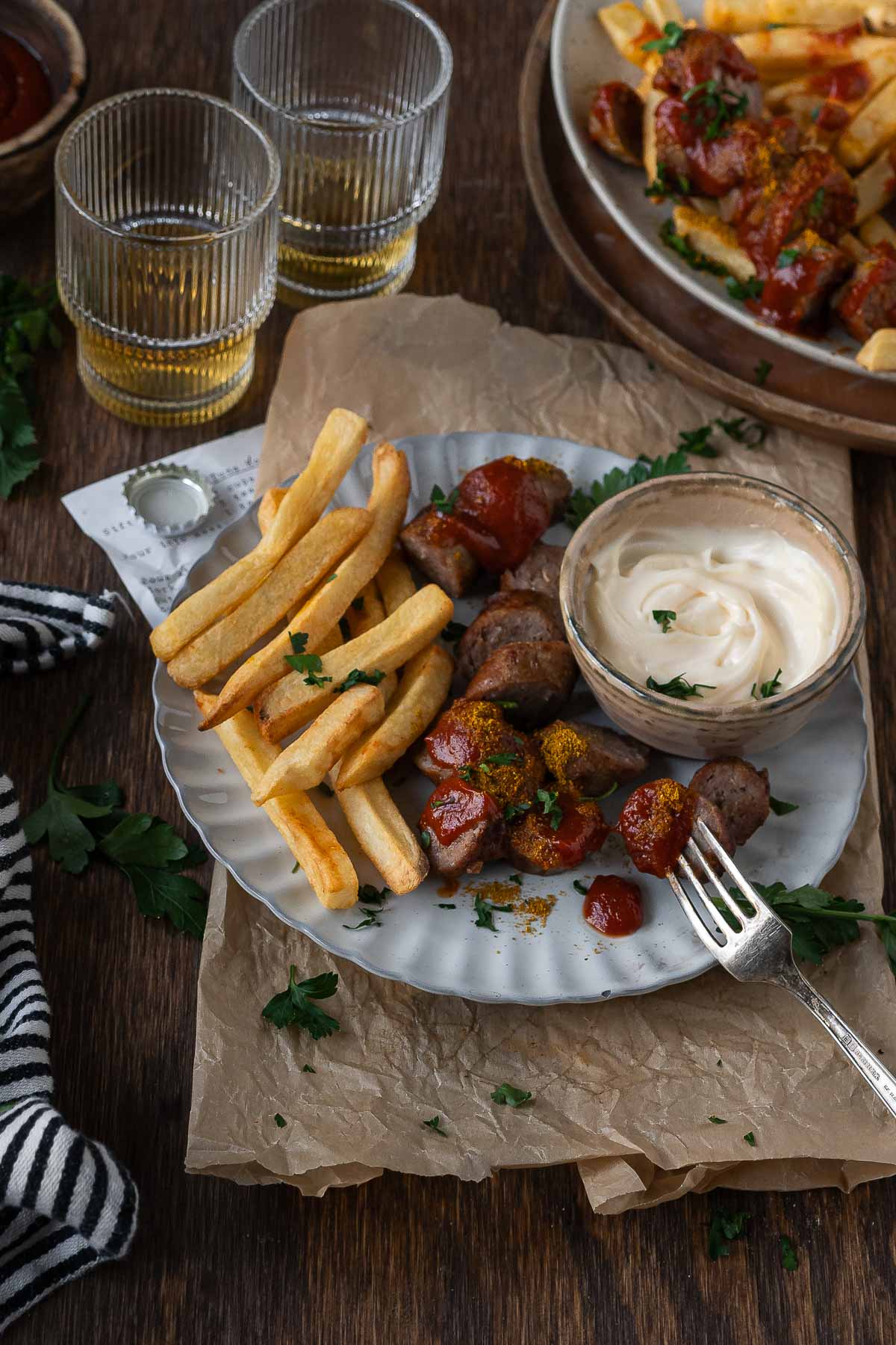 A plate of fries with sausage pieces topped with ketchup and herbs, served with a side of mayonnaise. Two glasses with drinks are in the background.