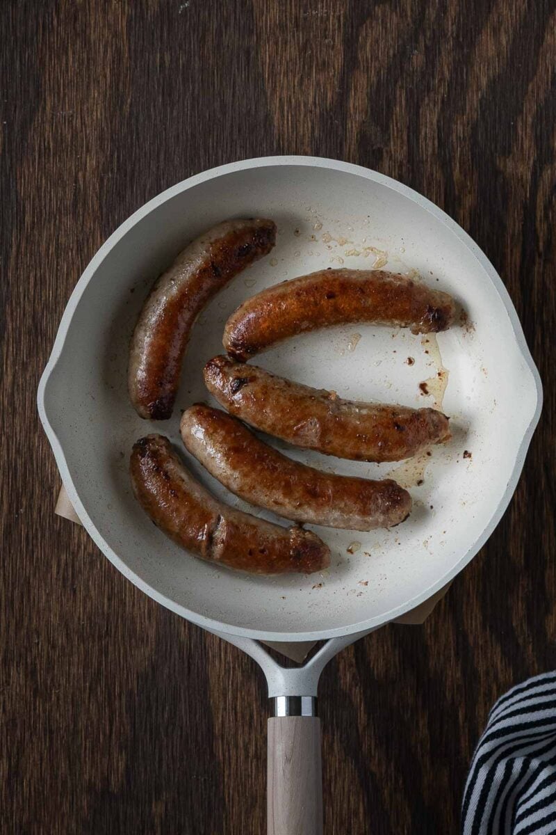 Five browned sausages cooking in a white frying pan on a dark wooden surface, with a striped cloth partially visible in the bottom right corner.