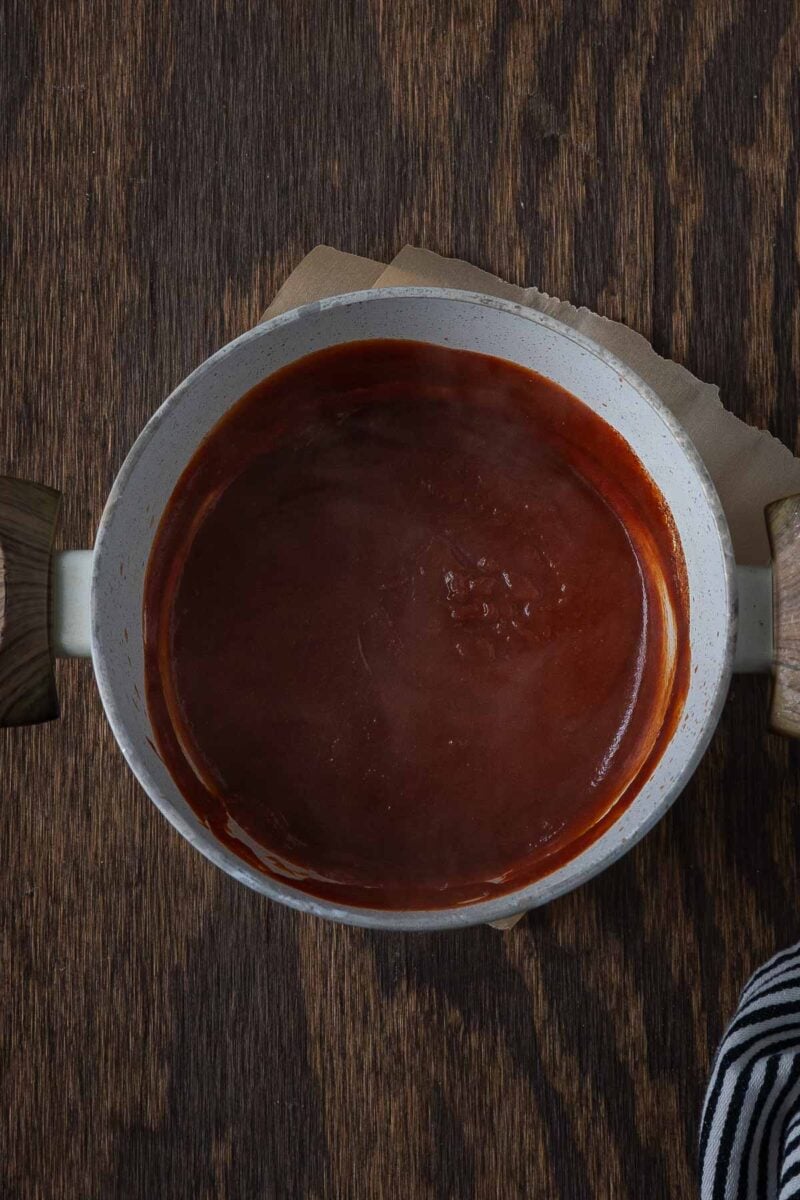 White saucepan filled with dark red sauce, placed on a wooden surface with handles visible on both sides.