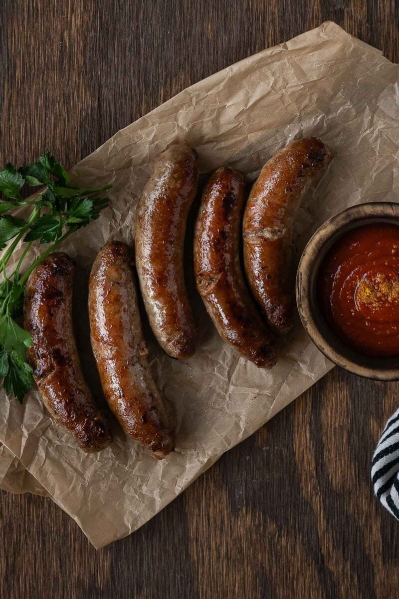 Five cooked sausages are arranged on parchment paper with parsley and a bowl of red sauce on a wooden table.