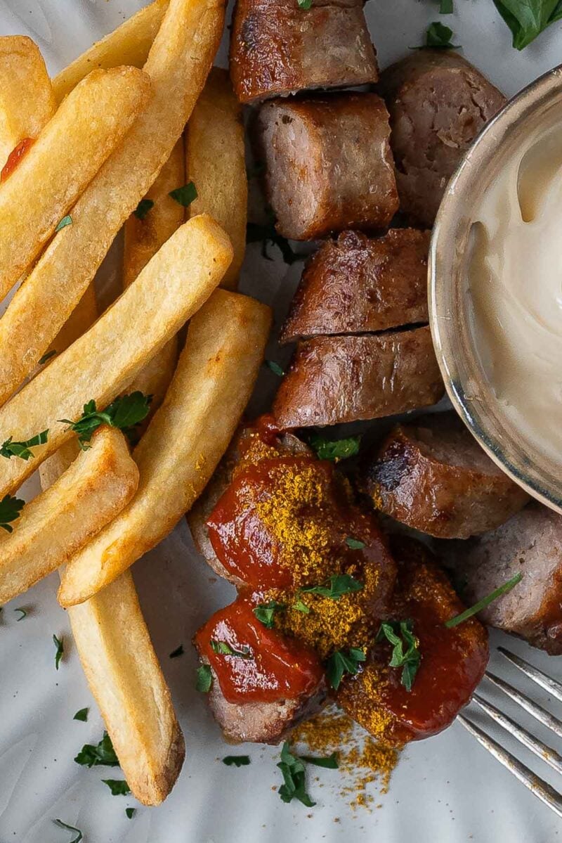 Plate with French fries, sliced sausage topped with ketchup and curry powder, chopped herbs, and a side of mayonnaise.