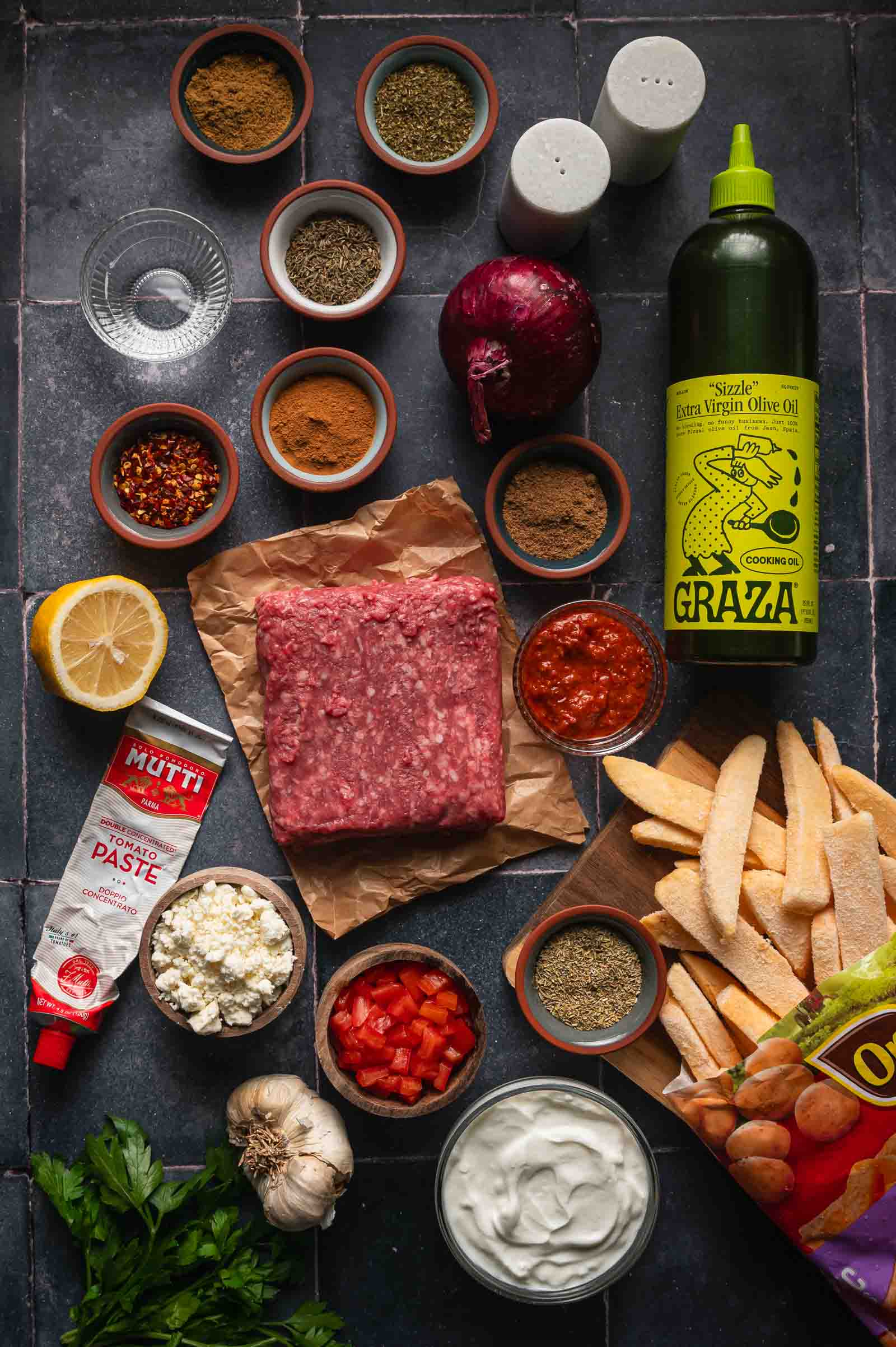 Ingredients for Greek feta fries: ground lamb, frozen steak fries, feta, Greek yogurt, harissa, tomato paste, red onion, garlic, lemon and the spice bowls.