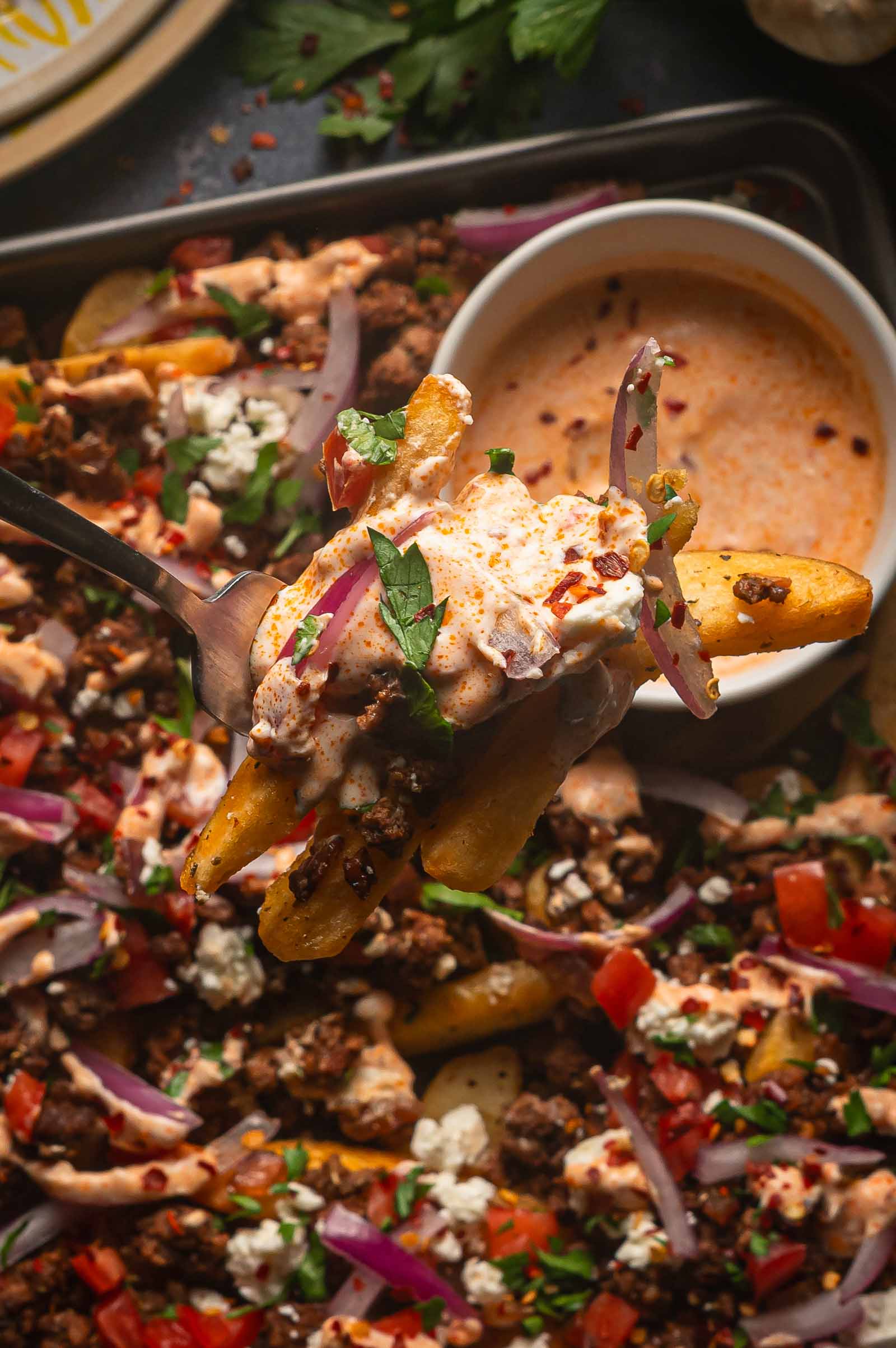 A forkful of Greek feta fries lifted from the pan, loaded with spiced ground lamb, feta, red onion and harissa yogurt sauce.
