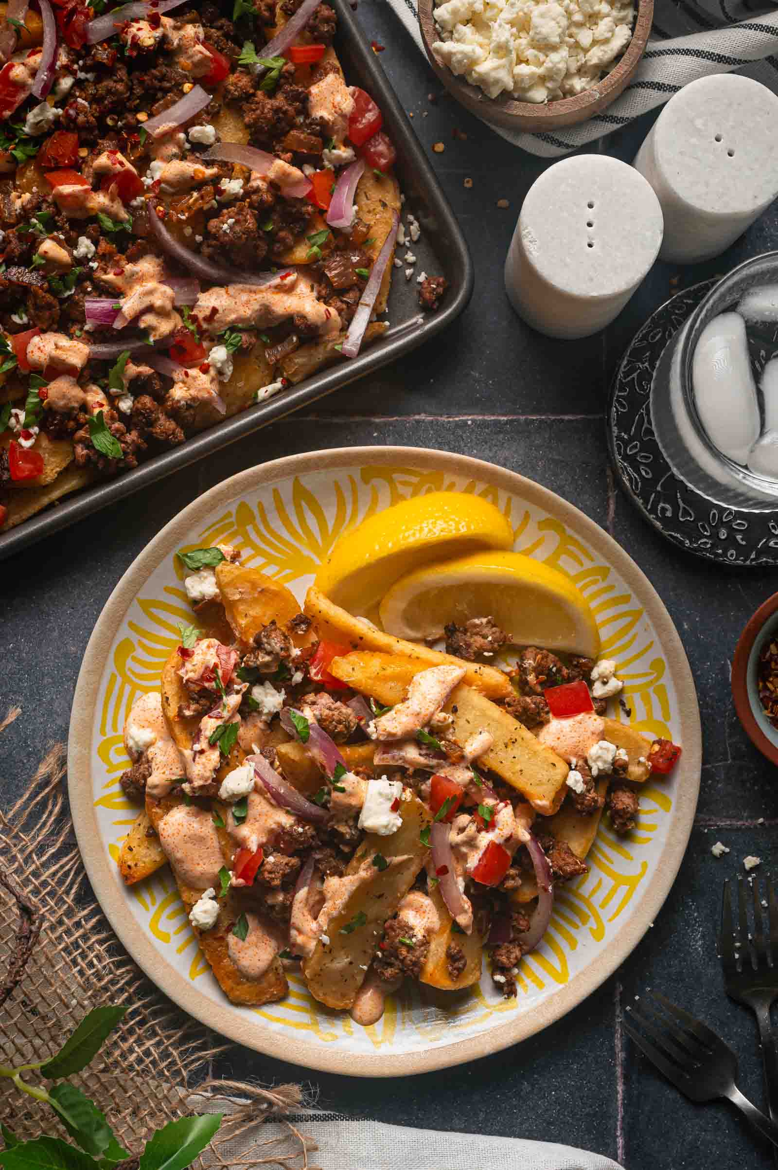A plate of loaded Greek-style fries with ground lamb, vegetables, feta cheese, and lemon slices, next to a tray of more fries, salt and pepper shakers, a bowl of feta, and a glass of ice water.