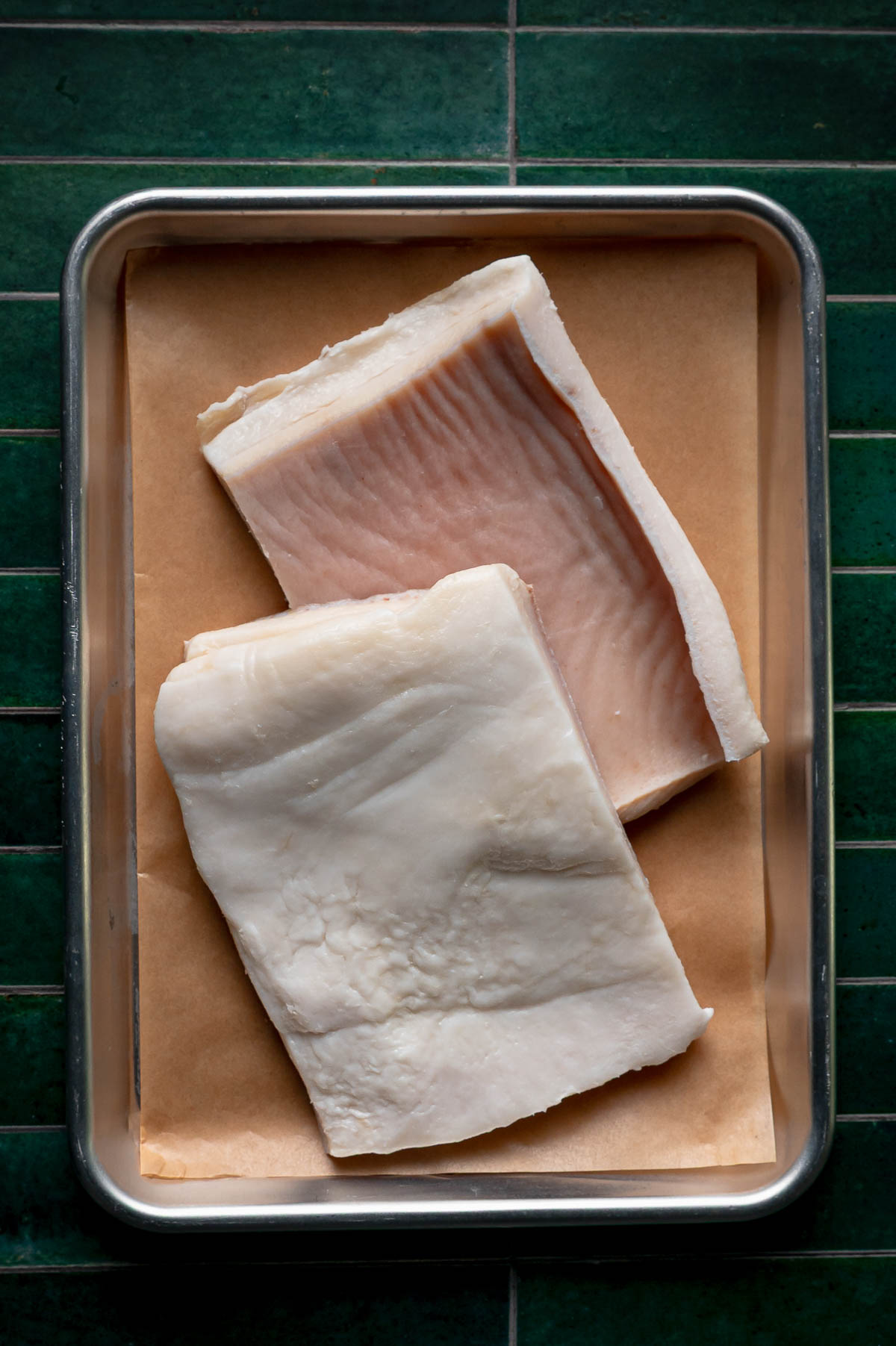 Two slabs of raw pork fat on parchment in a metal sheet pan, one still carrying its rind, ready to be cubed for rendering lard.