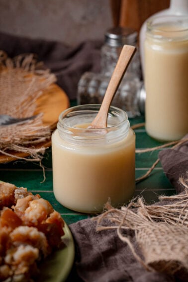 Two jars of finished slow cooker lard chilled firm, a wooden spoon standing in the open one and pork cracklings on a plate beside them.