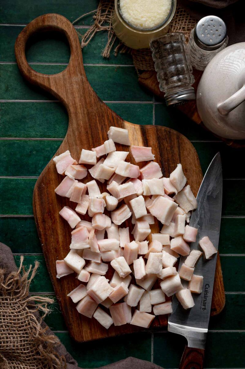Raw pork fat cut into rough half inch cubes on a wooden board with a chef's knife, sized evenly so every piece renders at the same rate.