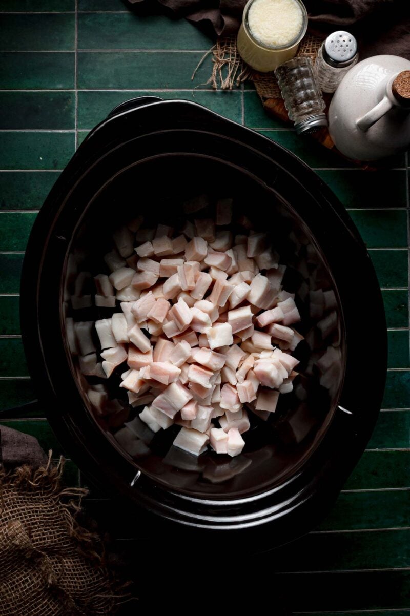Cubed pork fat spread in an even layer in a black slow cooker crock, the start of dry rendering lard with no water added.