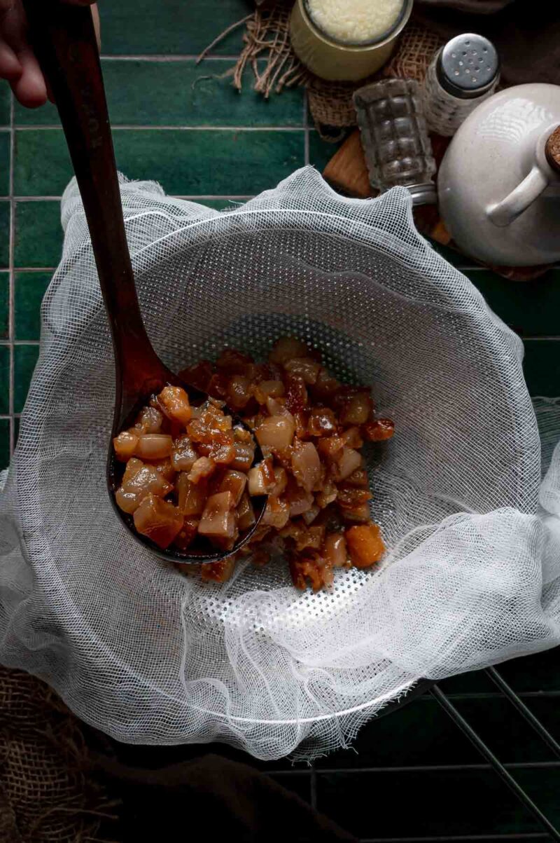 A wooden spoon lifting rendered pork solids into a cheesecloth lined fine mesh strainer to separate them from the liquid lard.