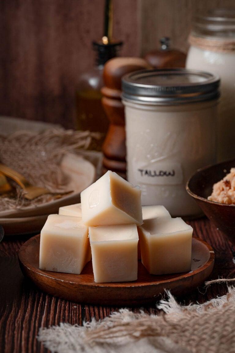 Cubes of set beef tallow stacked on a wooden plate beside the labeled jar and a bowl of rendered cracklings.