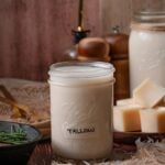 A mason jar of homemade beef tallow, cooled and set, on a rustic table with molded cubes of tallow and a cast iron pan of rosemary.