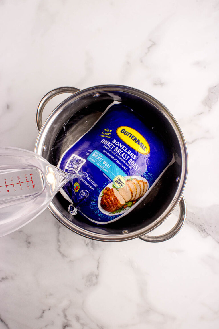 A packaged boneless turkey breast roast in a pot, with water being poured over it, on a marble countertop.