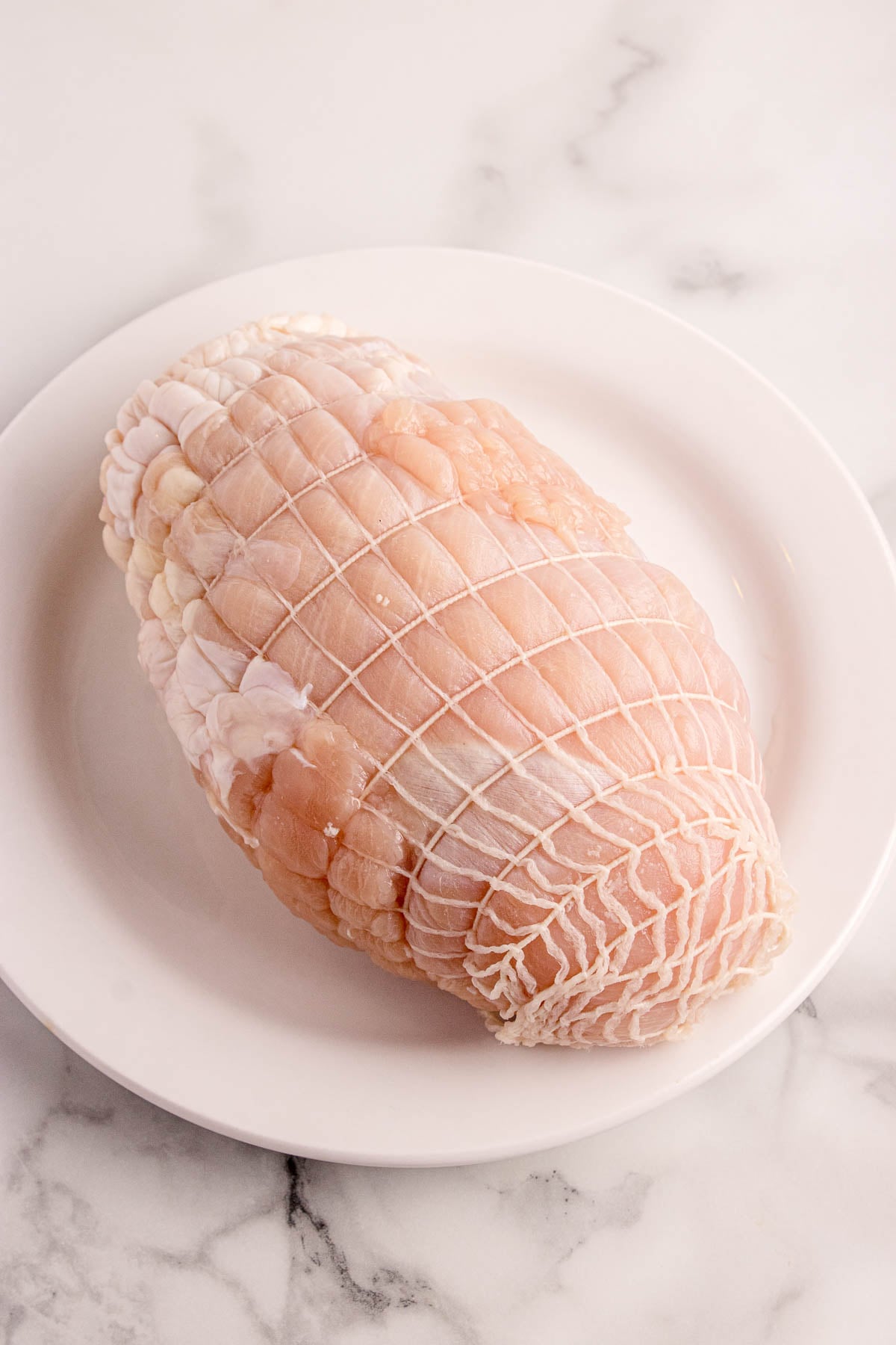 Raw turkey breast roast wrapped in butcher’s netting, placed on a white plate atop a marble surface.