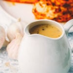 Make ahead turkey gravy in a white gravy boat on a lace cloth, with a head of garlic and a browned seasoned poultry breast behind it.