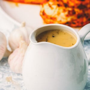 Make ahead turkey gravy in a white gravy boat on a lace cloth, with a head of garlic and a browned seasoned poultry breast behind it.