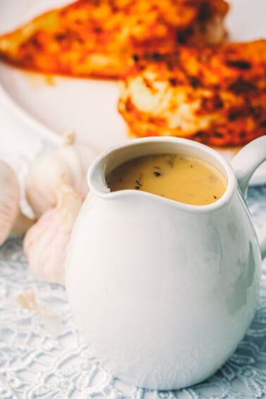 Make ahead turkey gravy in a white gravy boat on a lace cloth, with a head of garlic and a browned seasoned poultry breast behind it.
