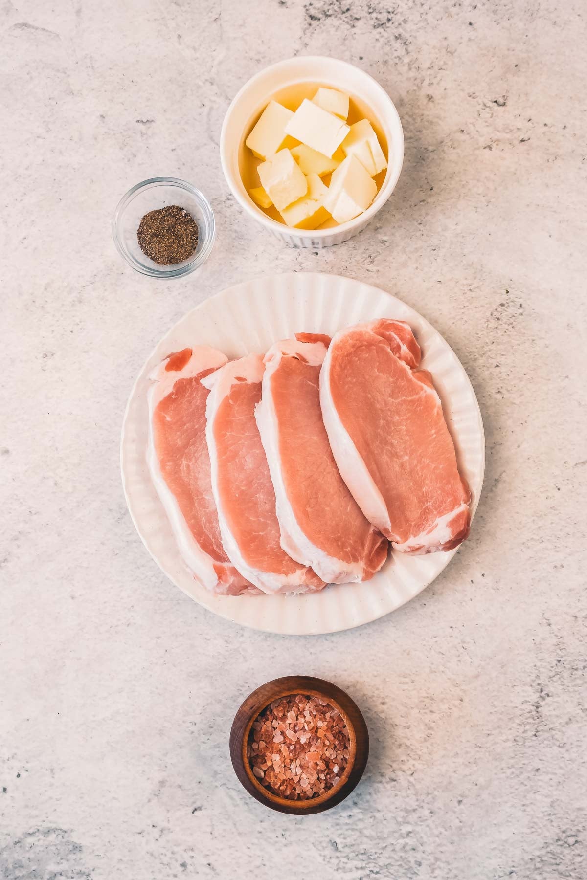 Four raw pork chops on a white plate, with bowls of cubed butter, ground black pepper, and pink Himalayan salt arranged around them on a light surface.