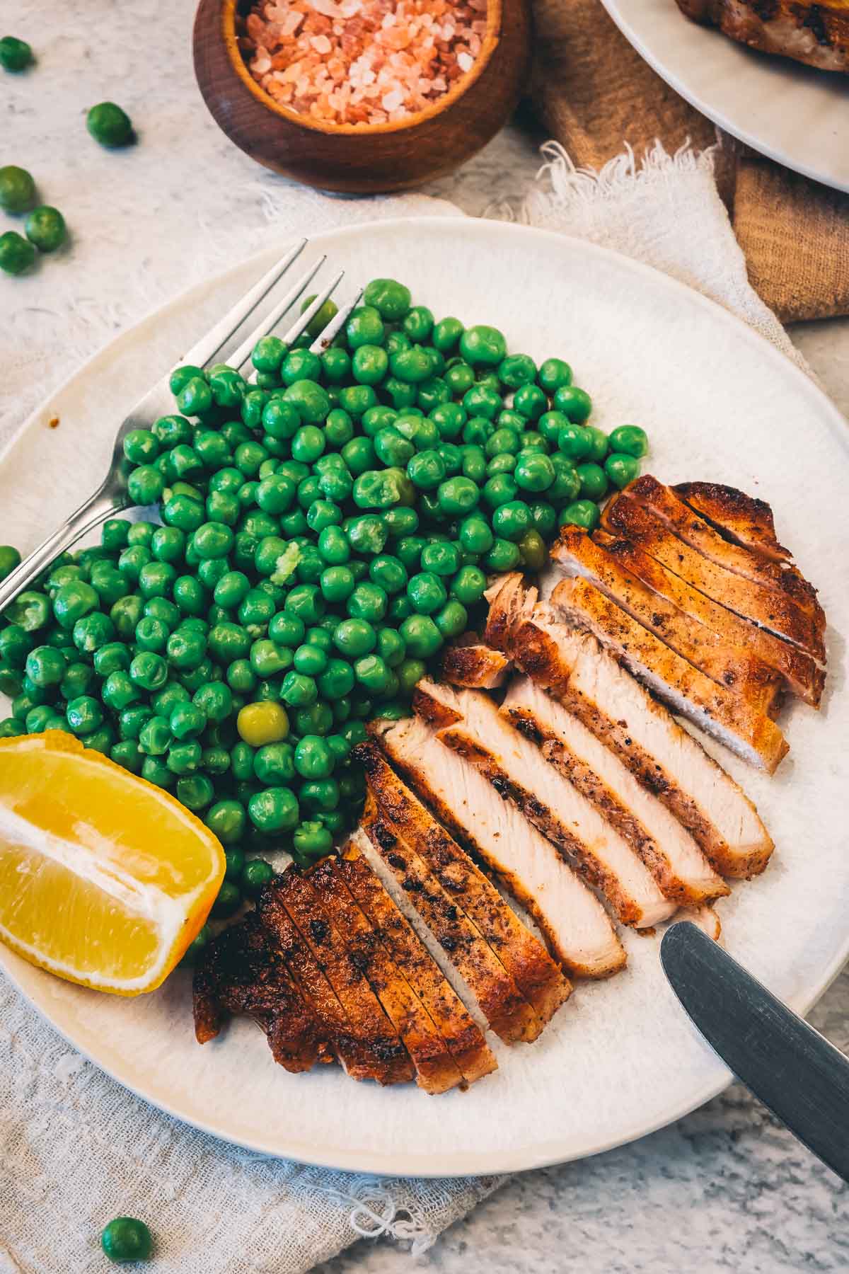 A sliced pan fried pork chop fanned beside green peas and a lemon wedge, showing the cut face inside the golden peppered crust.