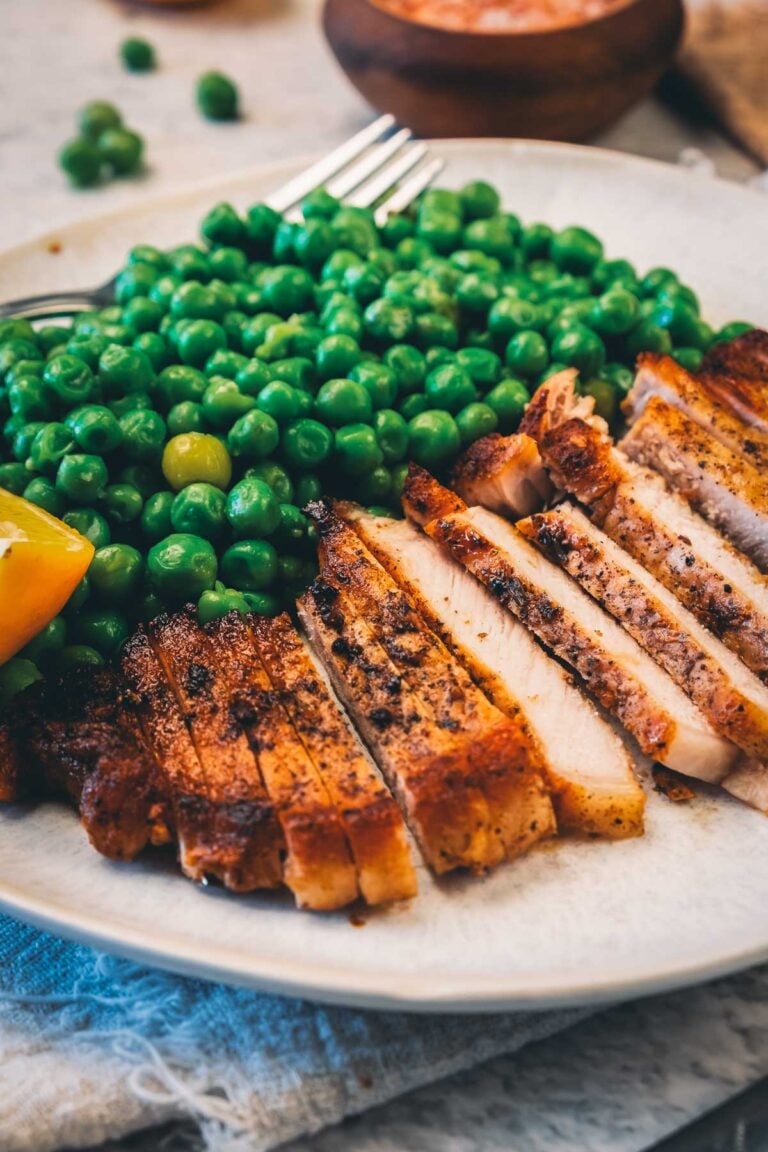 A sliced pan fried pork chop fanned across a plate of green peas with a lemon wedge, the peppered sear visible on every edge.