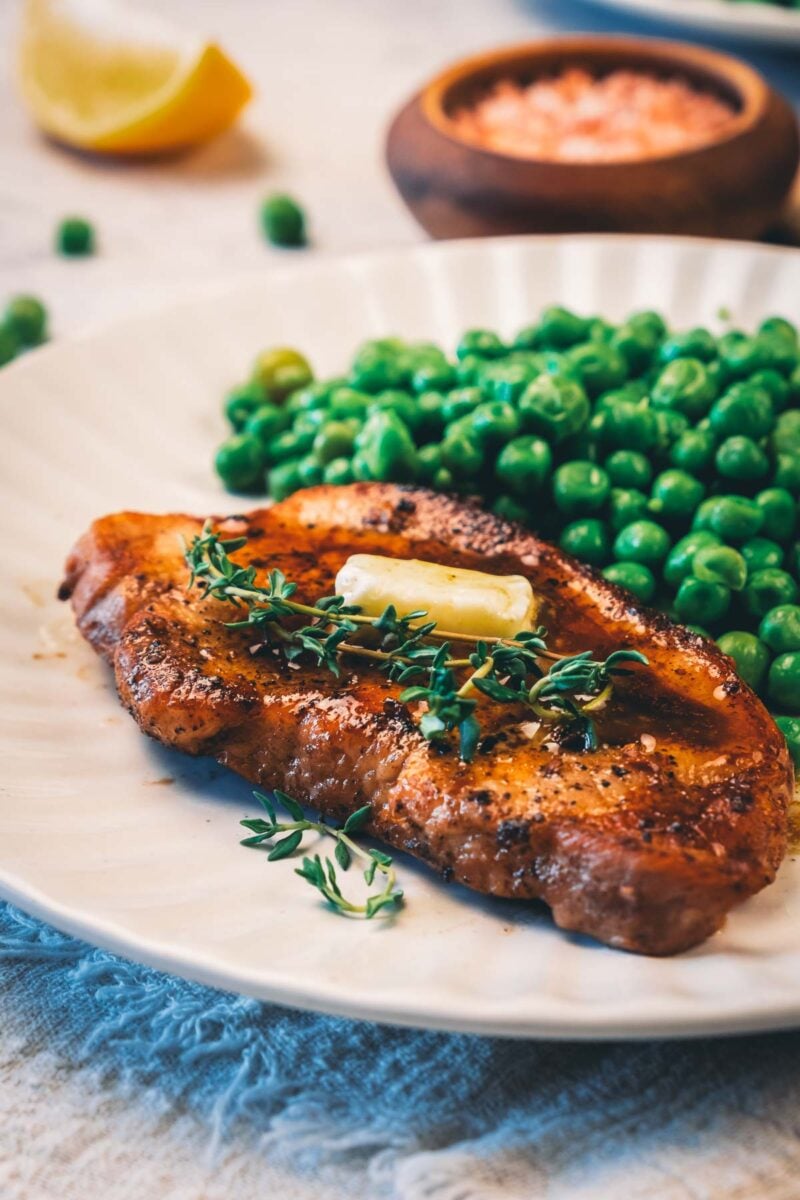 A pan fried pork chop topped with a pat of butter and fresh thyme sprigs, served with green peas on a white plate.