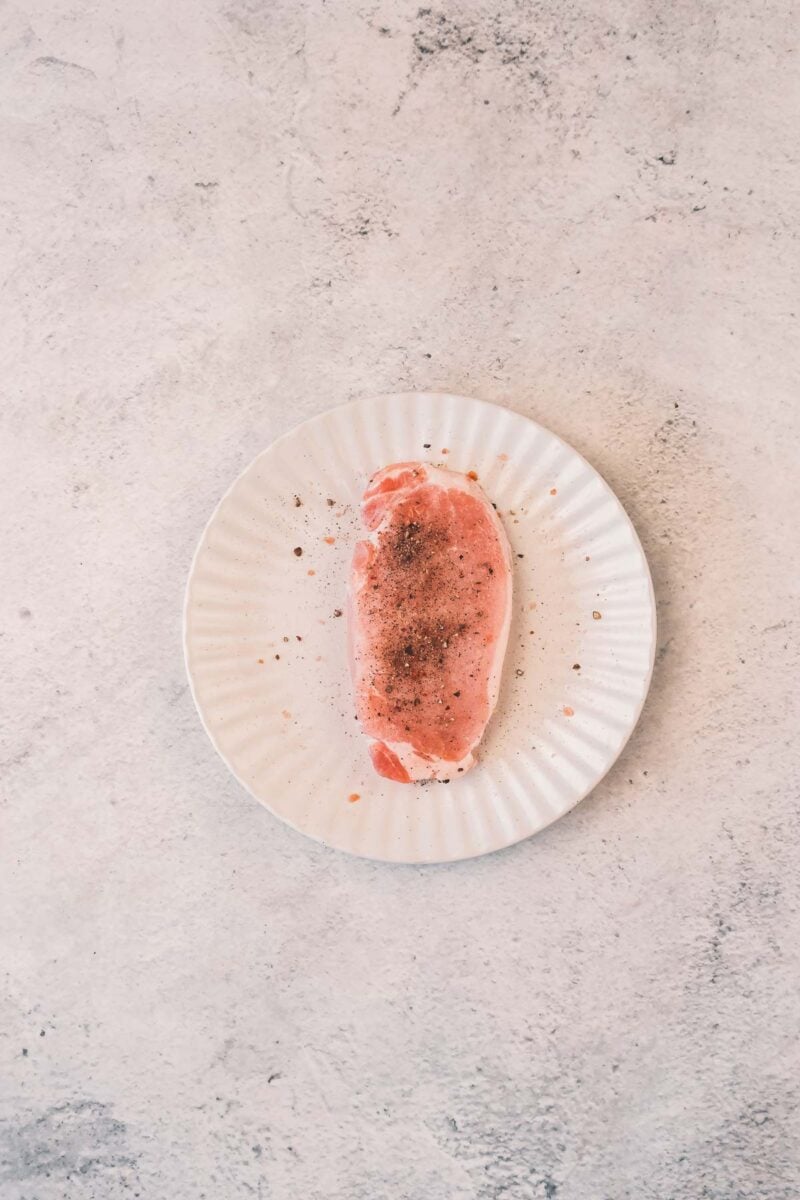 A raw boneless pork chop seasoned with pink salt and cracked black pepper on a white plate, ready for the skillet.