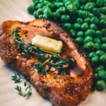 Close-up of a pat of butter and fresh thyme resting on a golden pan fried pork chop, with green peas behind.