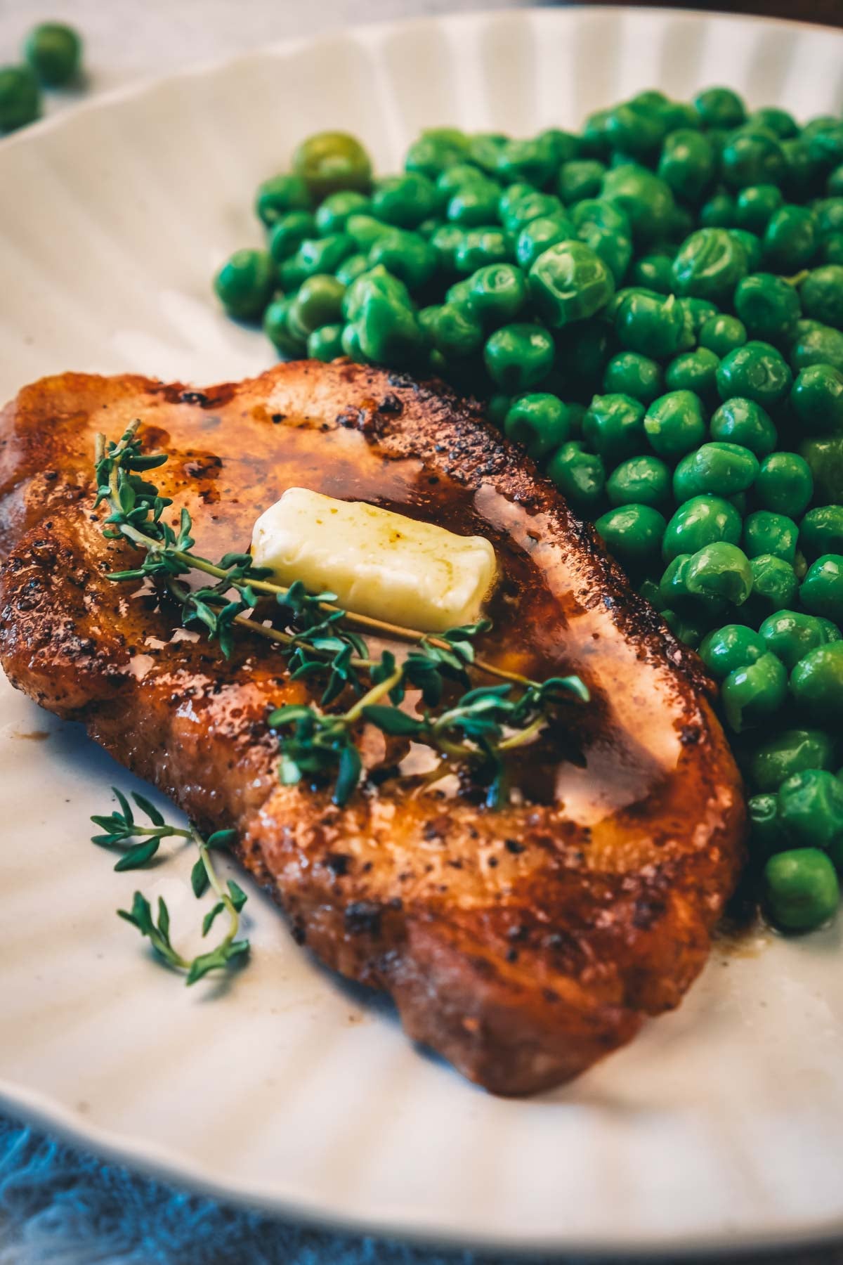 Close-up of a pat of butter and fresh thyme resting on a golden pan fried pork chop, with green peas behind.