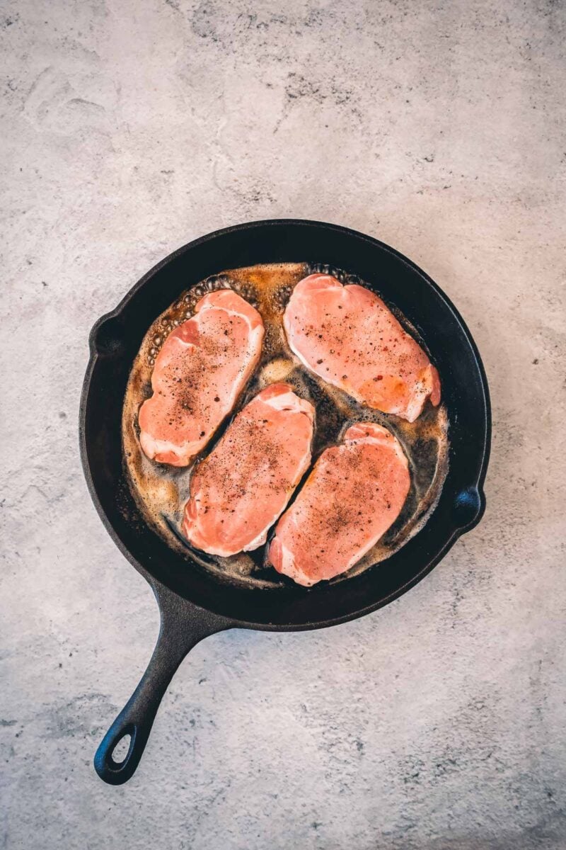 Four seasoned boneless pork chops just laid into a hot cast iron skillet, the first side starting to sear.