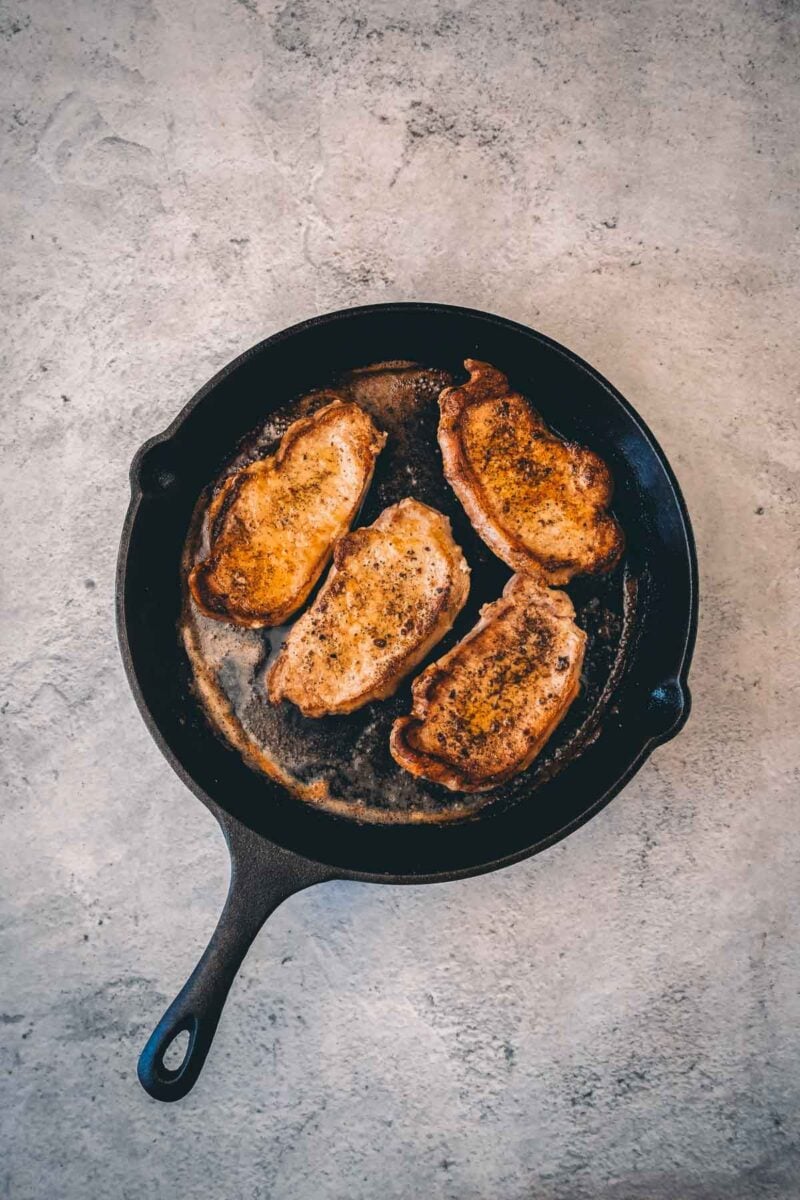Four pork chops flipped in the cast iron skillet, the seared side deep golden brown and crusted with black pepper.