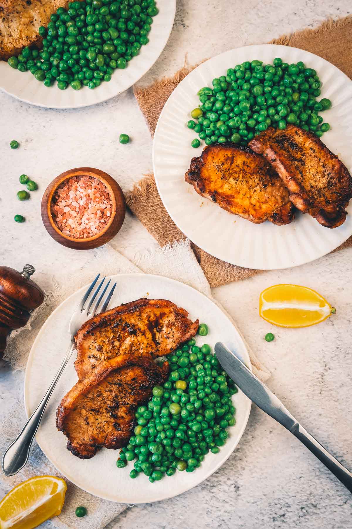 Two plates of pan fried pork chops and green peas from above, set with lemon wedges, a bowl of pink salt and a pepper mill.