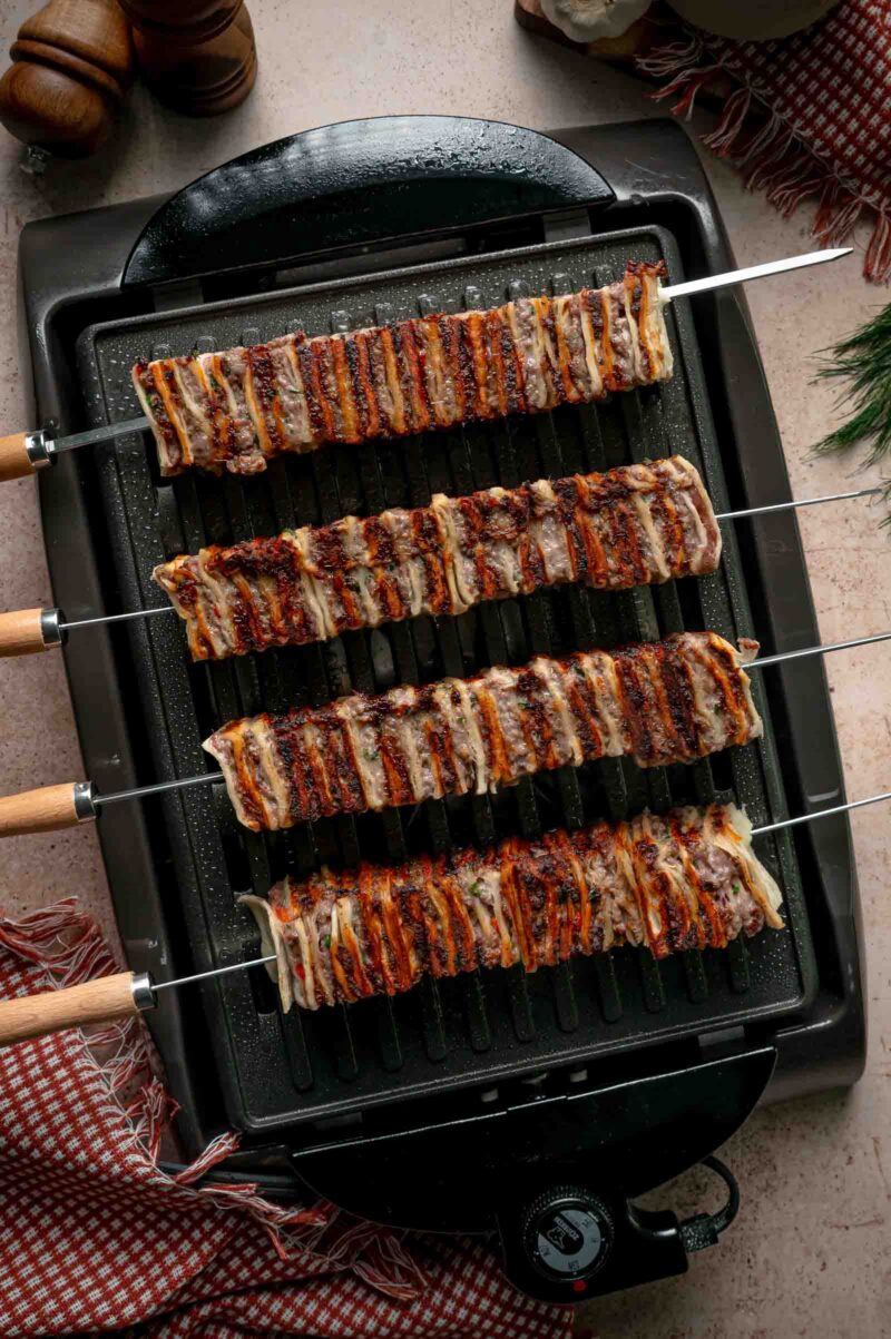 Four tortilla kebab skewers grilling on an indoor electric grill, the exposed tortilla edges charring dark.