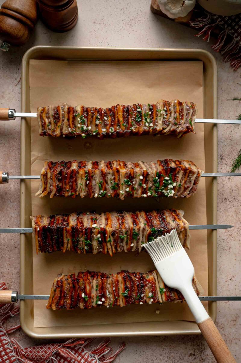 The grilled skewers on a parchment lined sheet pan being brushed with melted garlic butter and parsley.
