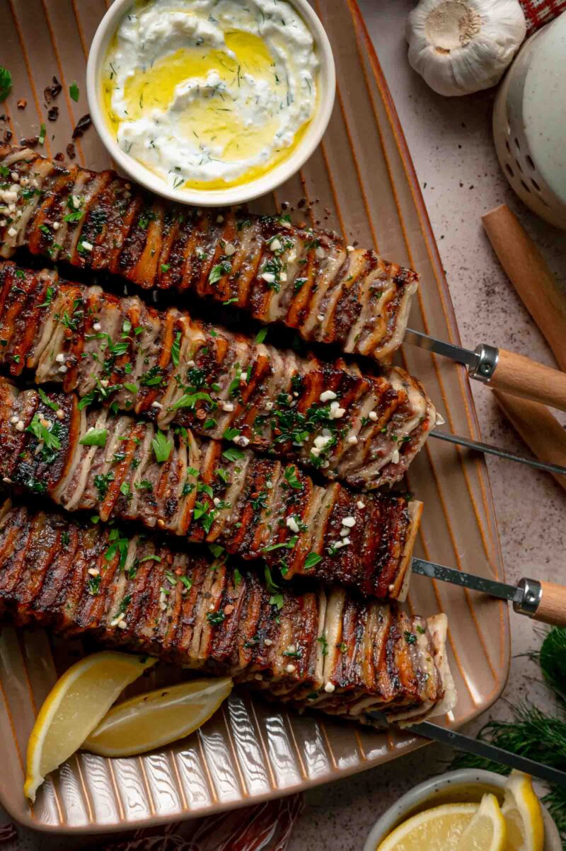 Tortilla kebab skewers crusted with garlic butter and parsley, lined up beside a bowl of haydari and lemon wedges.