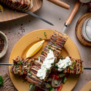Two tortilla kebab skewers crossed on a plate with dollops of haydari, a lemon wedge and chopped cucumber tomato salad.