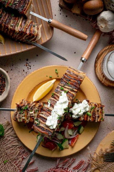 Two tortilla kebab skewers crossed on a plate with dollops of haydari, a lemon wedge and chopped cucumber tomato salad.